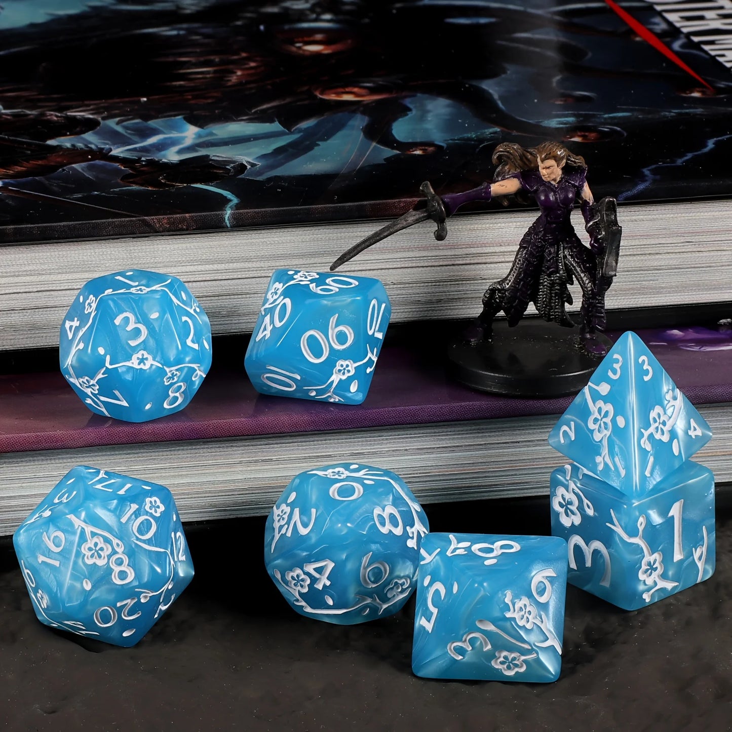 A set of blue polyhedral gaming dice with white engraved numbers and flower motifs, arranged on a dark tabletop in front of stacked books and a small fantasy figurine.