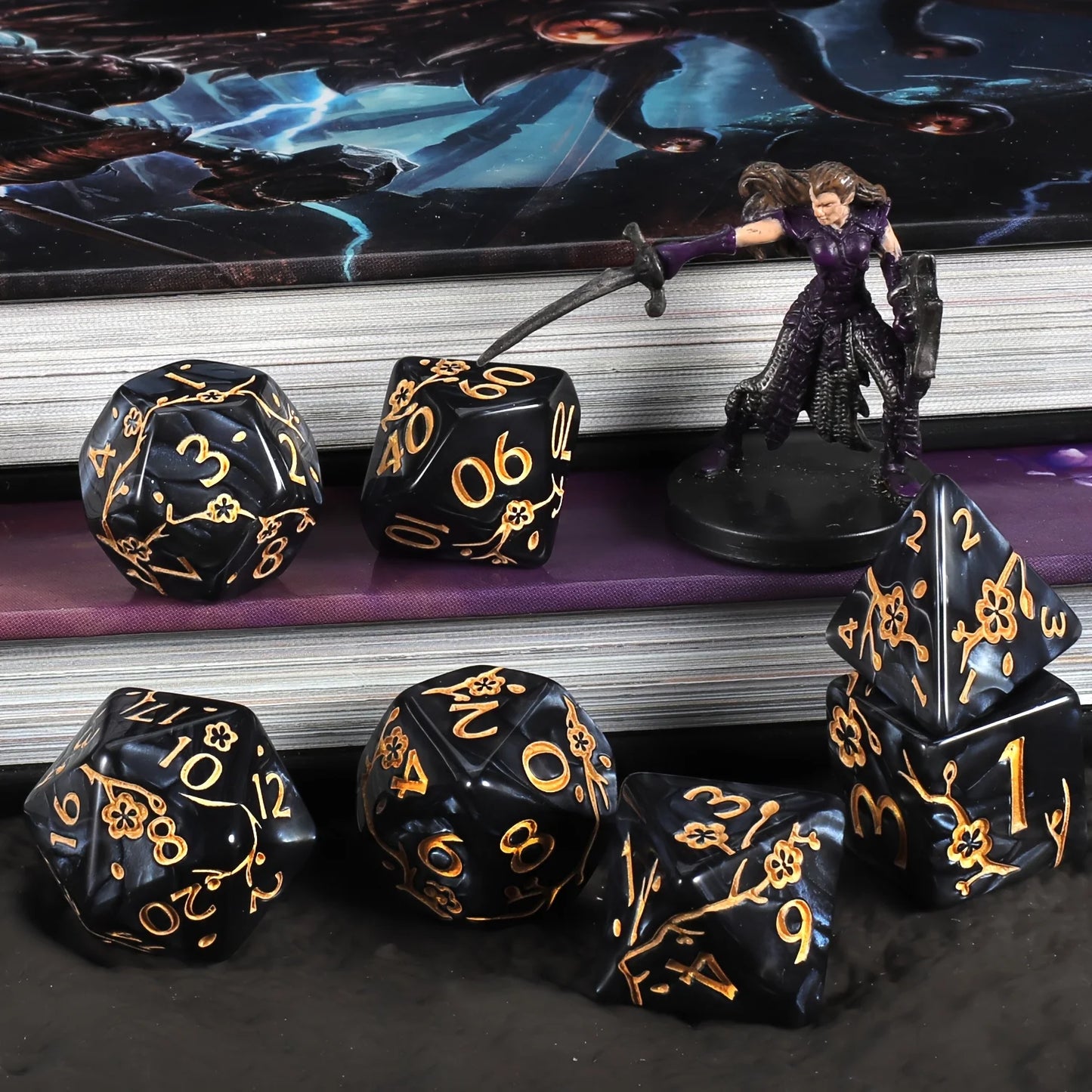 A set of black polyhedral tabletop gaming dice with gold numbers and ornate gold flower motifs, arranged on dark surfaces in front of stacked books. A small fantasy figurine wearing a purple outfit stands on one of the dice.