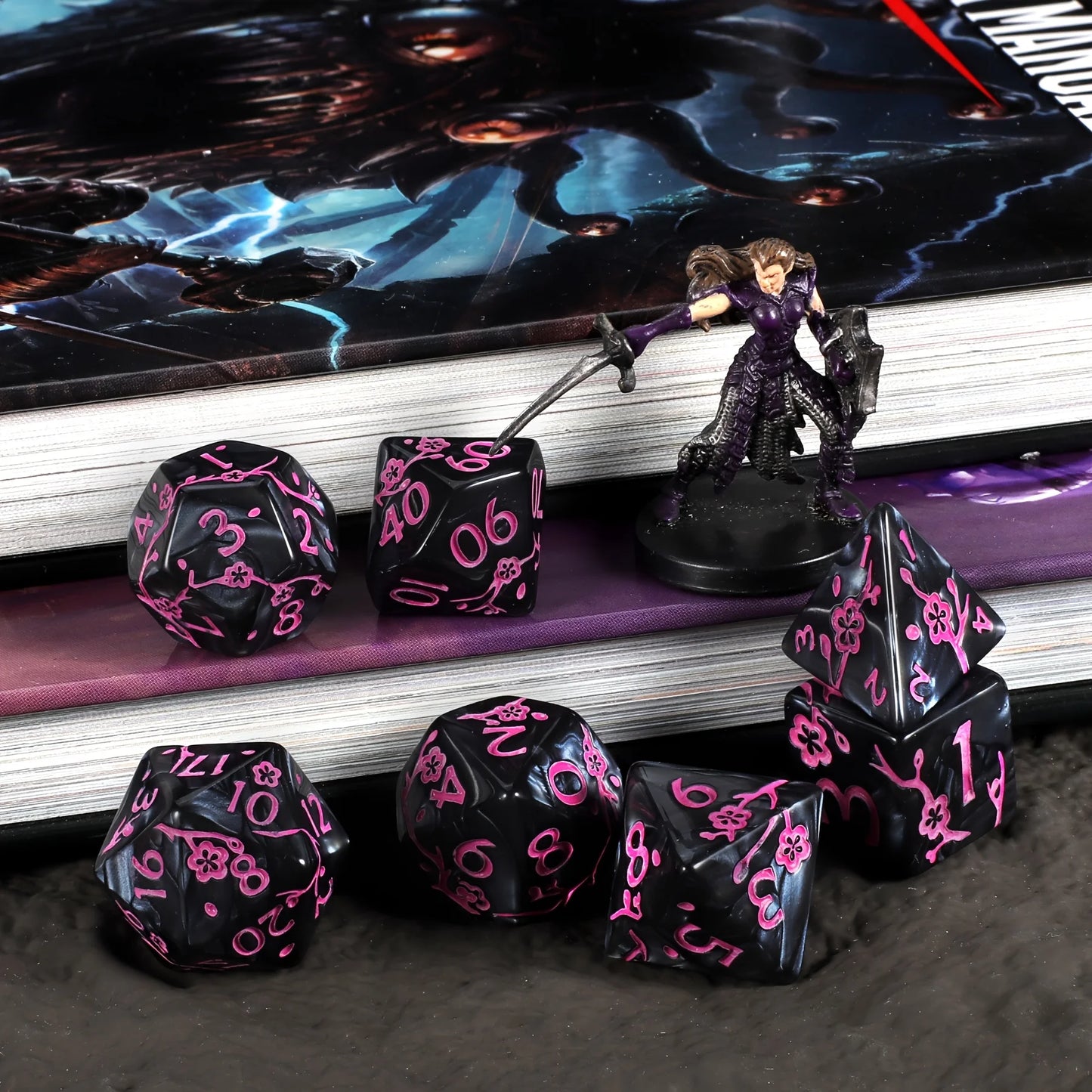 A set of black polyhedral gaming dice with pink flower motifs, arranged on a dark surface in front of a book and a small fantasy figurine.