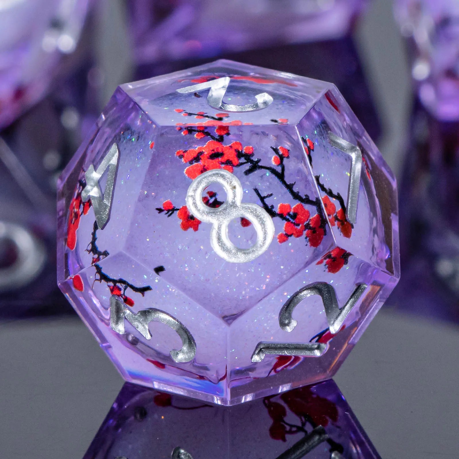 A translucent purple 20-sided d20 die featuring silver-colored numbers and red cherry blossom branches with white flowers embedded inside, displayed on a reflective surface.