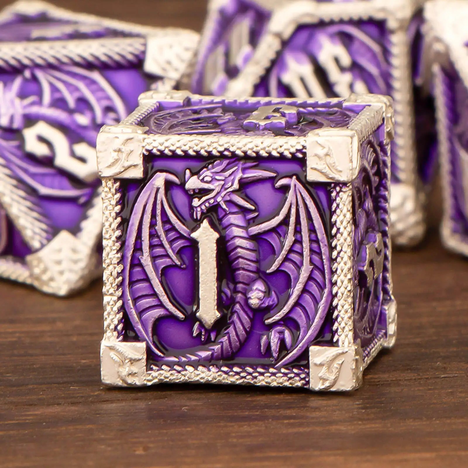 A purple and silver fantasy dice box featuring an embossed dragon design on the front, displayed on a wooden surface with other matching dice blurred in the background.