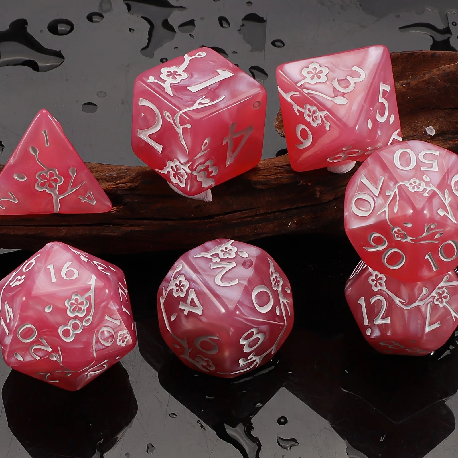 A set of pink polyhedral dice with white engraved numbers and floral designs, arranged on a dark reflective surface with a wooden prop.