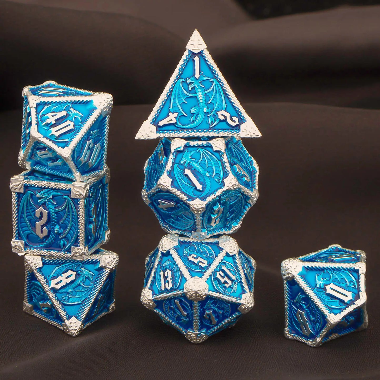 A set of metal blue and silver polyhedral gaming dice displayed on a dark brown fabric background. The set includes a d20, d10, d8, d6, d4, and d100, all featuring a textured blue surface with embossed silver numbers and white decorative borders.