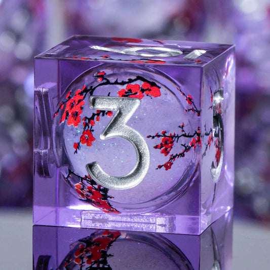 A transparent purple acrylic dice cube showing a silver number 3 with red cherry blossom branches on the inner face, photographed close-up against a blurred purple background.