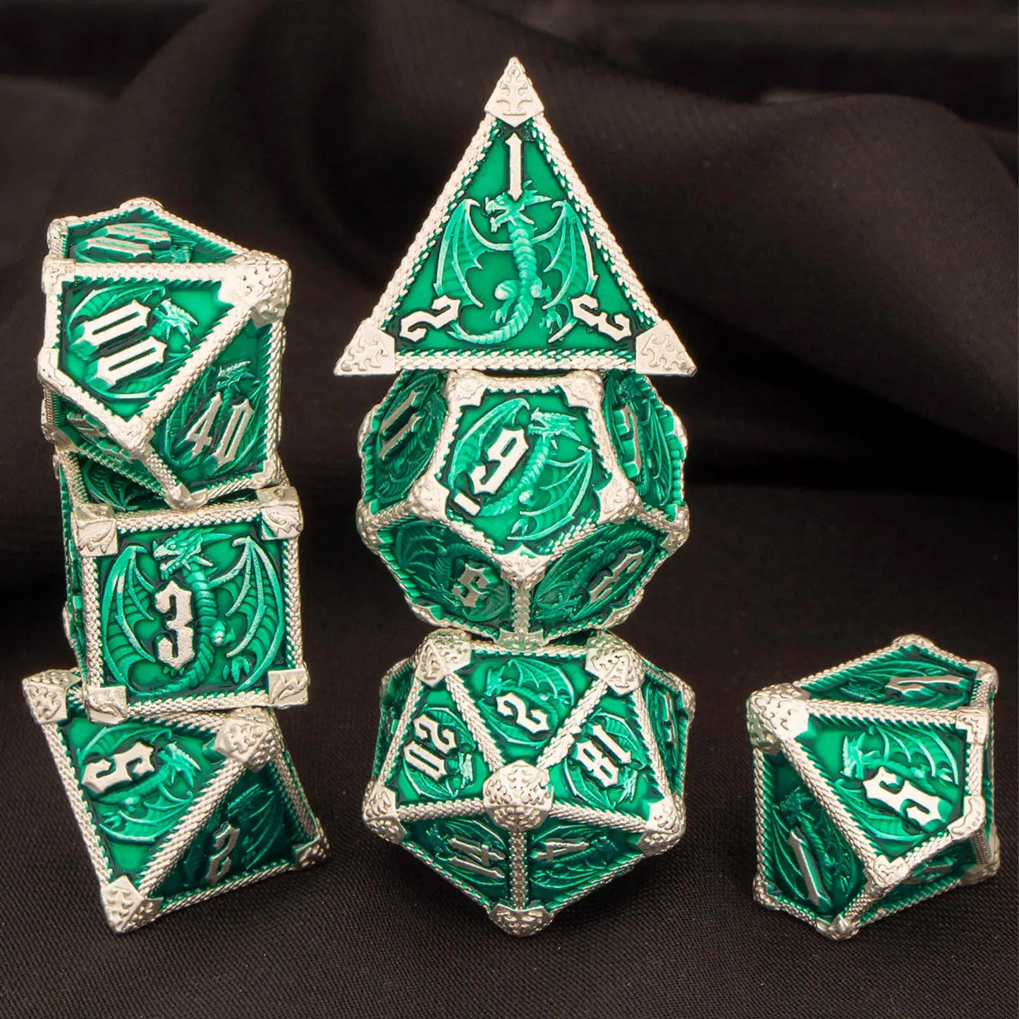 A set of metal green and silver dragon-themed polyhedral dice, including a d4, d6, d8, d10, d%, d12, and d20, arranged on a dark draped fabric background.