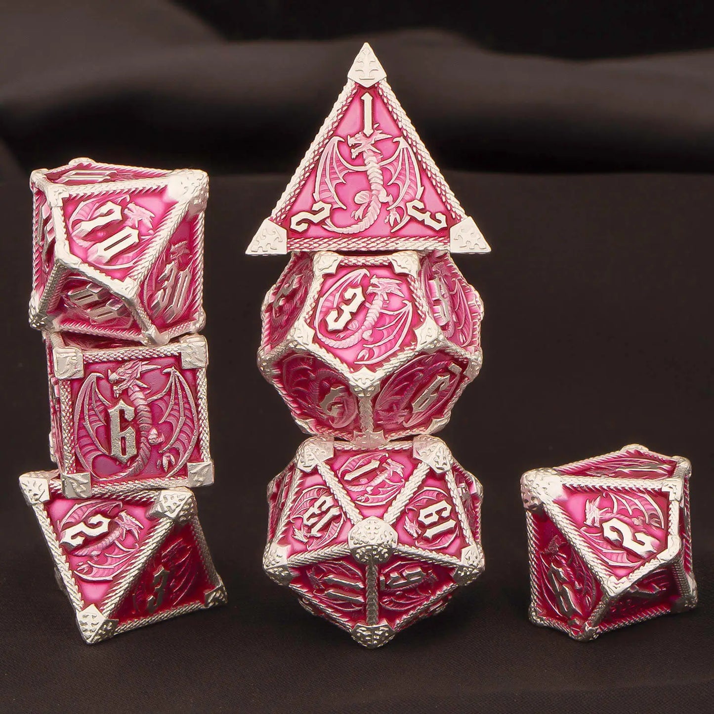 A set of metal pink and silver fantasy-themed polyhedral dice displayed on a dark fabric background. The set includes a d4, d6, d8, d10, d%, d12, and d20, all featuring detailed silver dragon engravings and numbering.