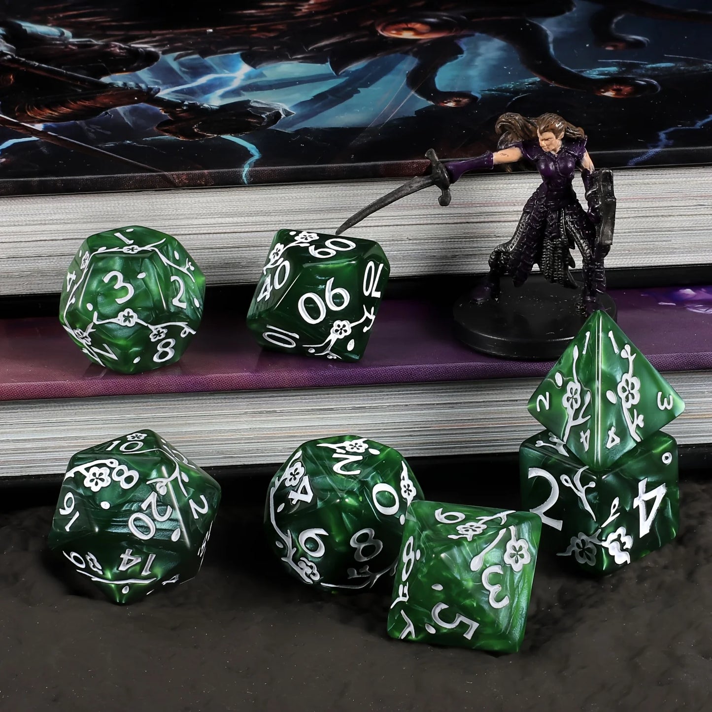A set of green polyhedral gaming dice with white numbers and flower motifs, arranged on a dark tabletop in front of stacked books and a fantasy figurine.