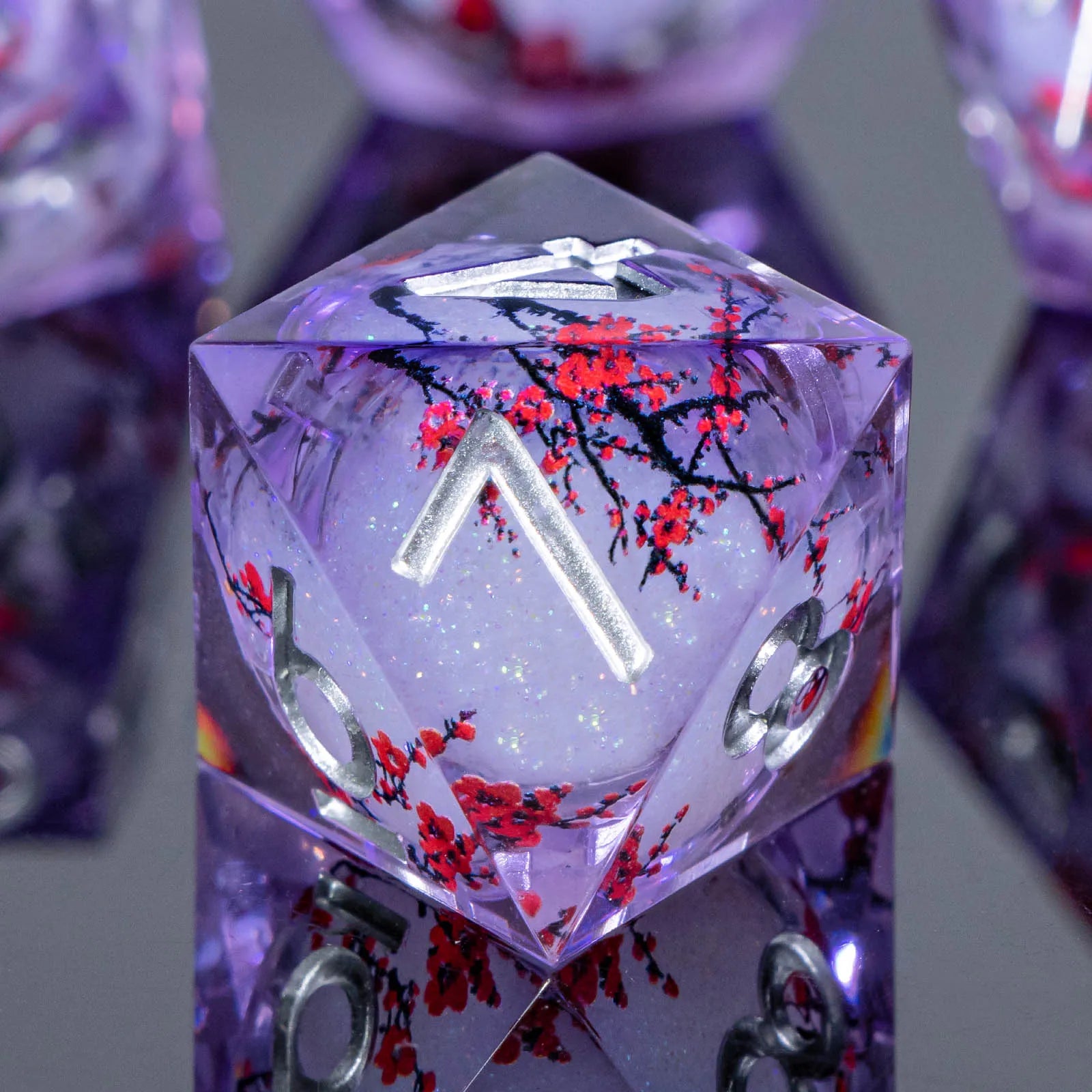 A close-up of a translucent purple polyhedral die showing a red cherry blossom pattern with silver-colored numbers, set against a blurred background of similar dice.