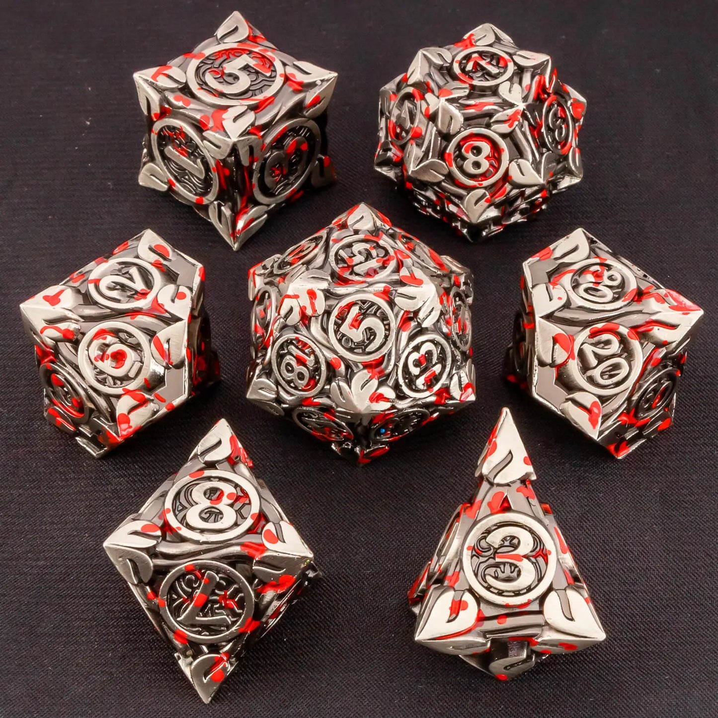 A set of ornate silver metal polyhedral gaming dice with red and black floral engravings, arranged on a dark textured fabric background.
