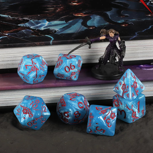 A set of light blue polyhedral gaming dice with red numbering and red flower symbols, arranged on and in front of a stack of books with a fantasy character figurine and a game mat in the background.