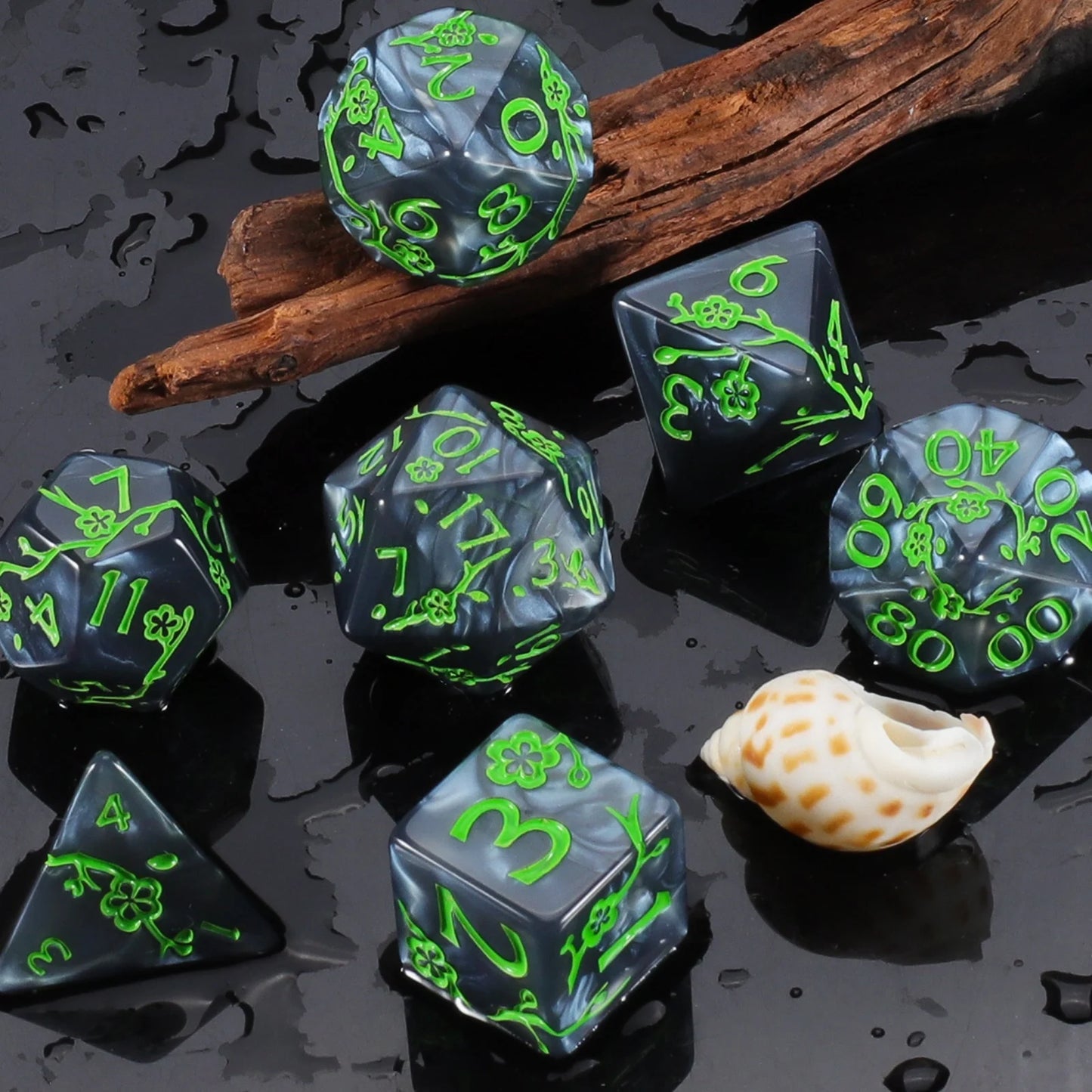 A set of dark gray polyhedral gaming dice with bright green engraved numbers and flower symbols, arranged on a glossy wet surface with a piece of driftwood and a small shell prop.