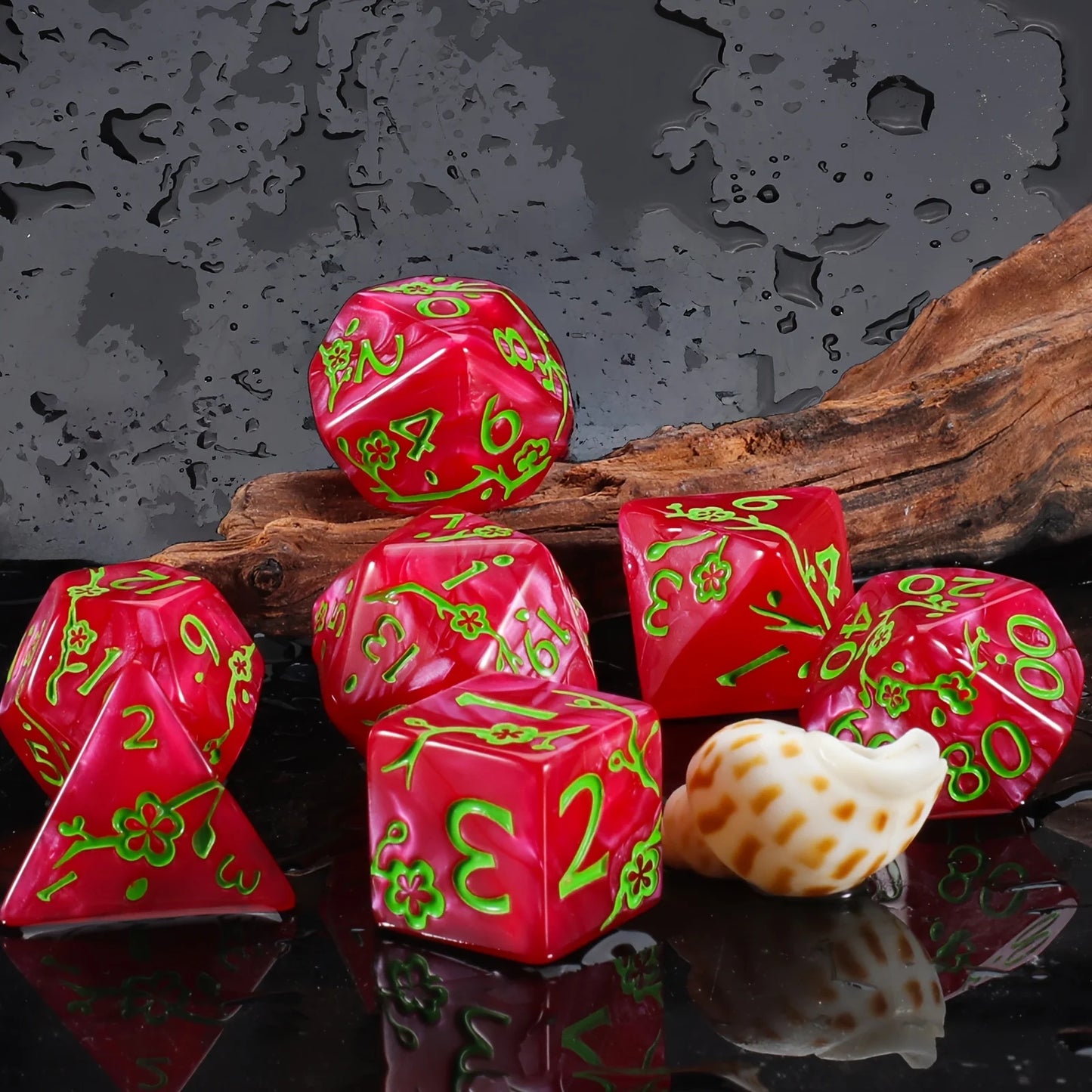 A set of red polyhedral dice with bright green numbering and floral accents, arranged on a dark reflective surface with a piece of driftwood and a small seashell prop against a wet, dark gray background.