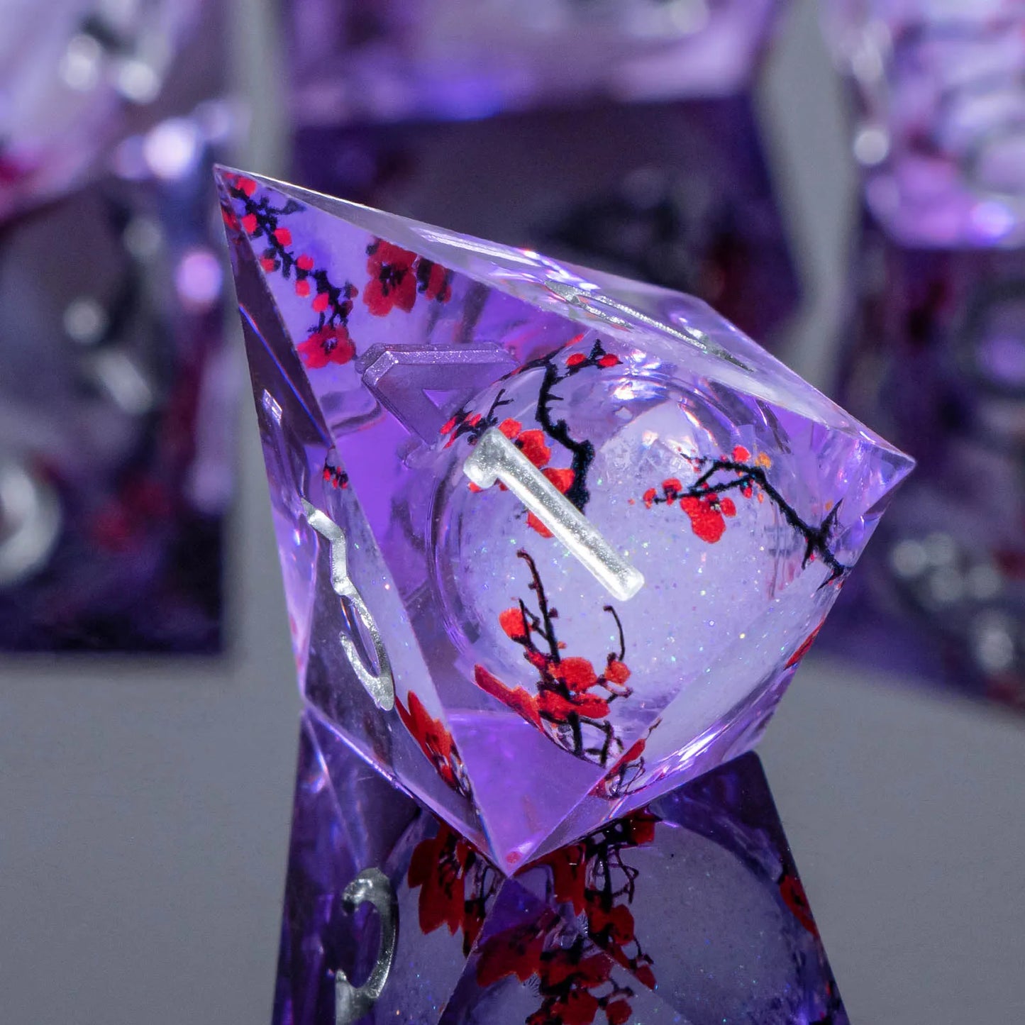A close-up of a faceted transparent purple polyhedral die with red and black floral inclusions, standing on a matching purple display base against a softly blurred background.