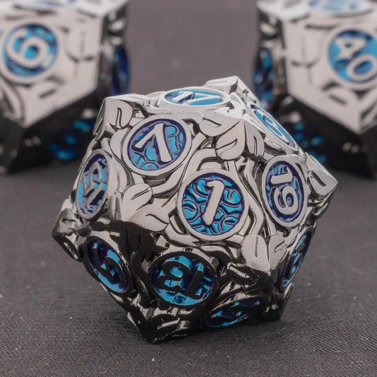 A metal 20-sided die (d20) from a polyhedral gaming set, featuring a metallic silver and black leaf pattern with vibrant blue circular inserts displaying white numbers. Two similar dice are blurred in the background on a dark gray textured surface.