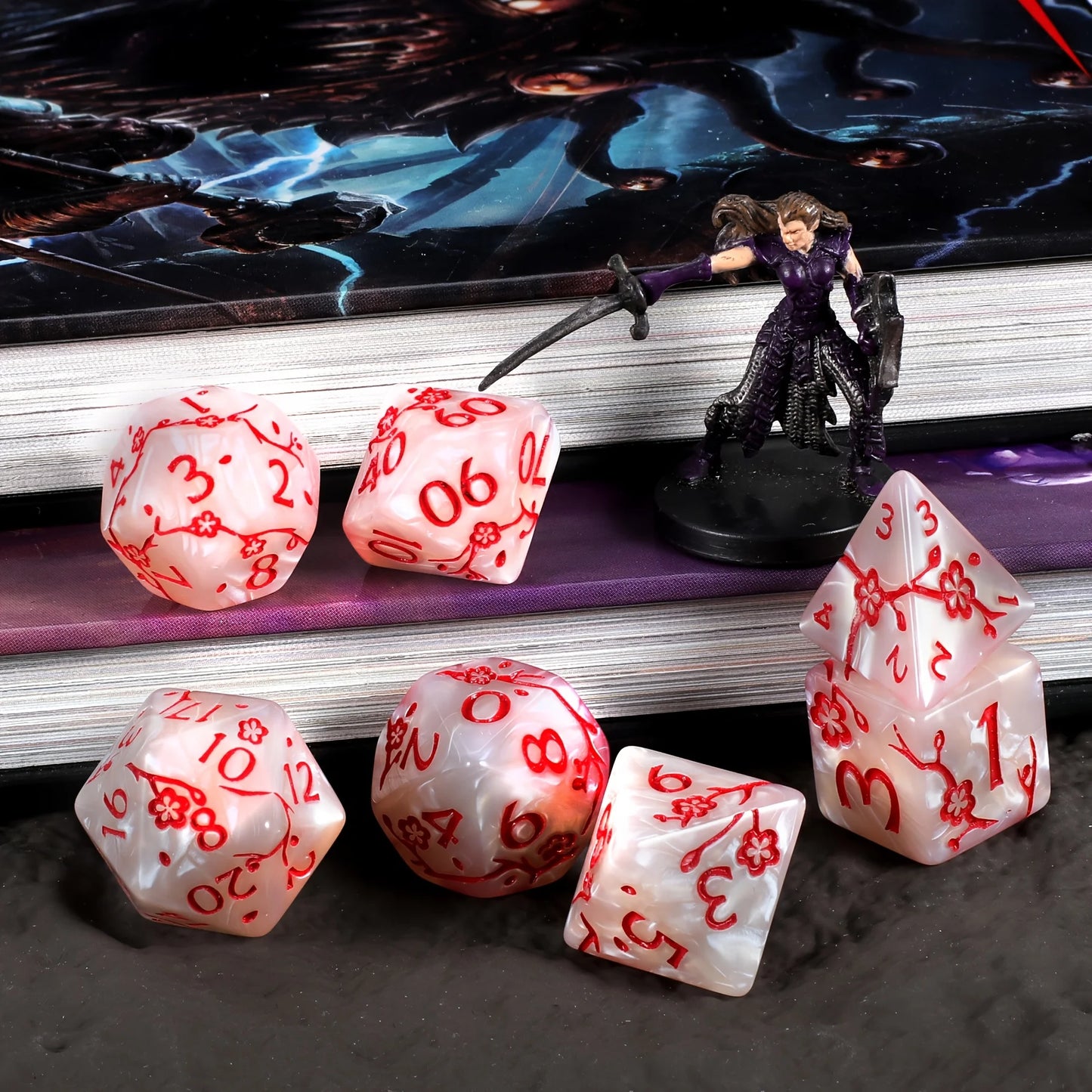 A set of white polyhedral gaming dice with red flower symbols and numbers, arranged on a dark surface in front of stacked books and a small fantasy figurine.