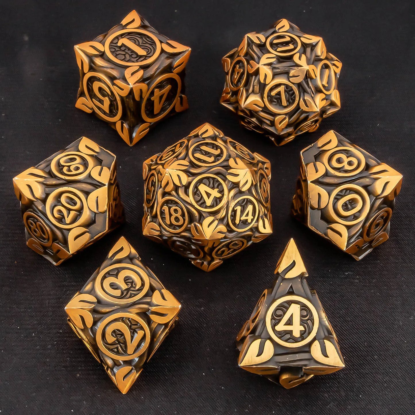 A set of seven metal fantasy-themed polyhedral dice with a dark metallic bronze and gold leaf pattern, arranged on a dark textured background.