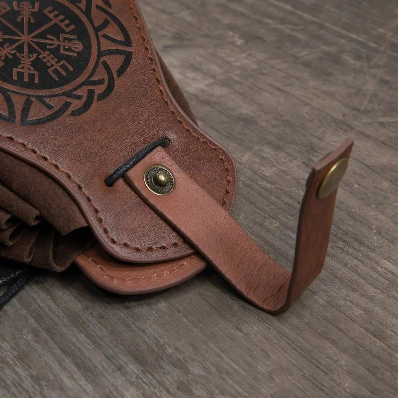 A close-up of a brown leather accessory, likely a belt or strap component, featuring a black Norse-style compass engraving, a metal snap button, and a brass-toned rivet, resting on a gray wood-textured surface.
