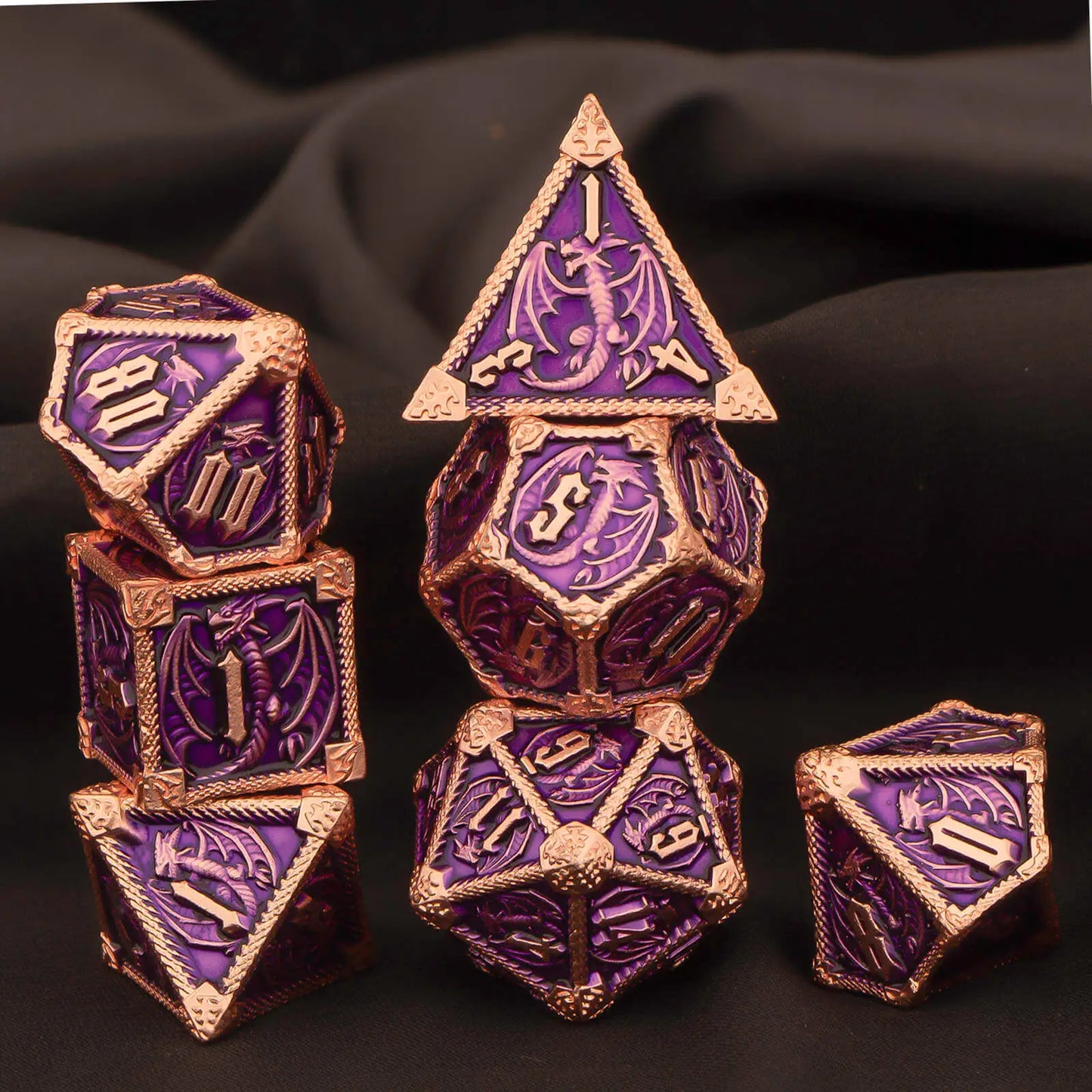 A set of metal purple and rose gold fantasy gaming dice with dragon motifs, arranged on a dark draped fabric background.