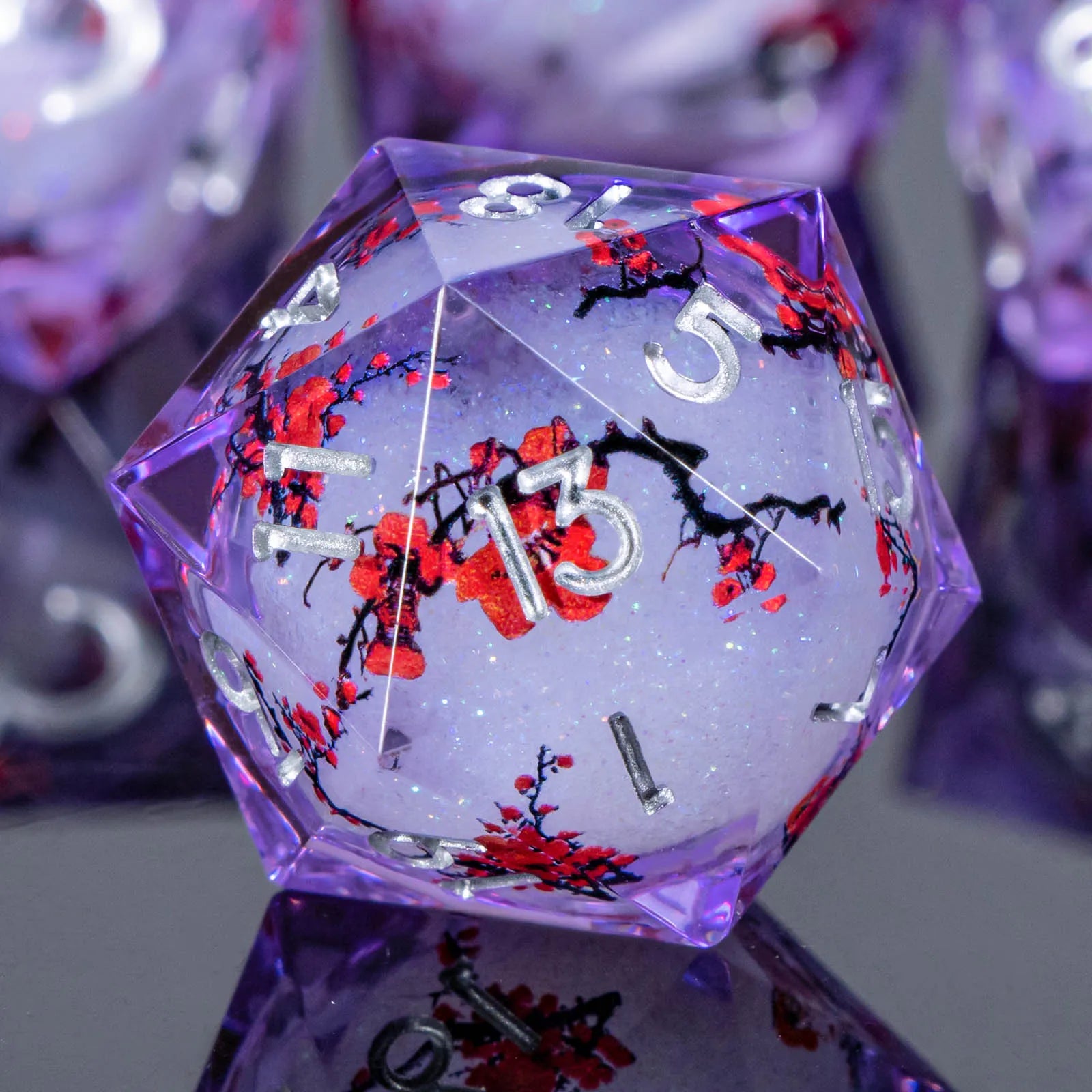 A translucent purple polyhedral die, likely a d20, filled with glitter and decorated with red cherry blossoms and black branches, shown in sharp focus against a blurred purple background.