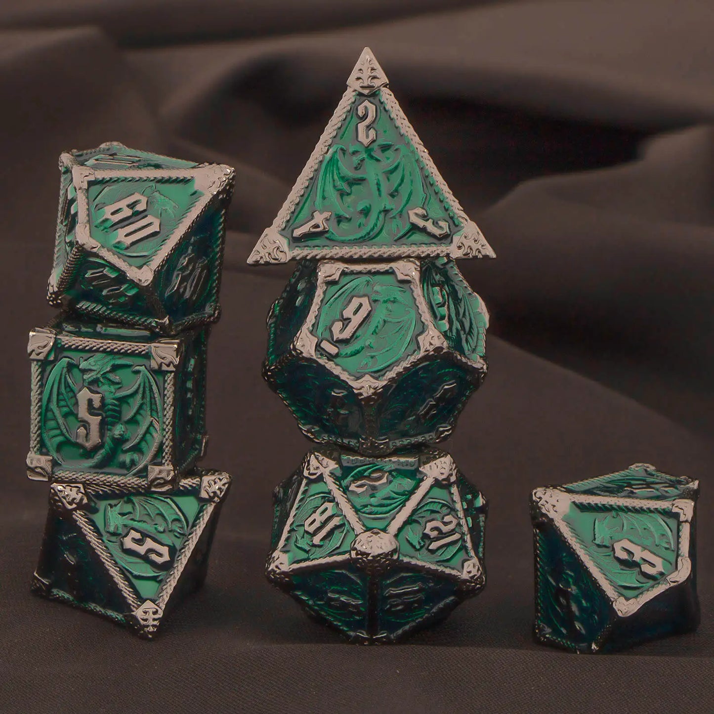 A set of green and dark silver metal polyhedral gaming dice arranged on a dark draped fabric background. Includes a d4, d6, d8, d10, d%, d12, and d20.